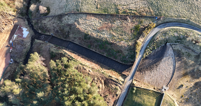Aerial view of a curved road passing through a rural landscape with patches of grass, trees, and a small building. A bridge crosses a narrow stream.